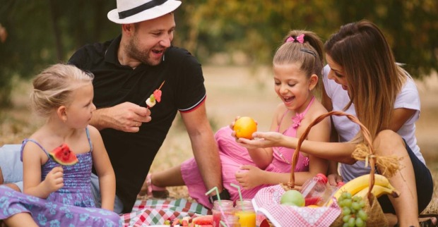 Eine fröhliche Familie beim Picknick im Freien. Zwei Kinder und ihre Eltern sitzen auf einer Decke, essen frisches Obst und lachen miteinander. Der Vater trägt einen weißen Hut und hält einen Obstspieß, während die Mutter und eine Tochter eine Orange betrachten. Im Vordergrund steht ein Picknickkorb mit Bananen, Trauben und Getränken. Die Szene findet an einem sonnigen Tag im Grünen statt.