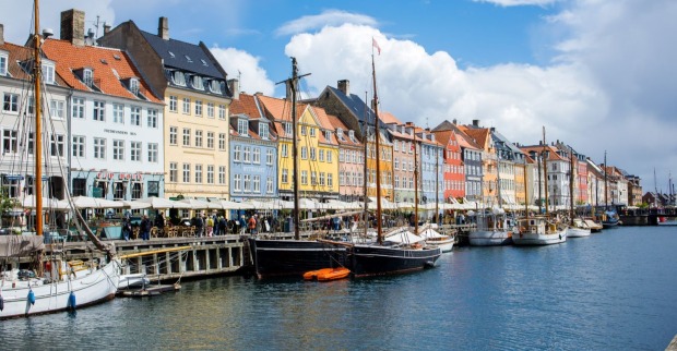 Bunte Giebelhäuser und historische Segelboote am Hafenkanal Nyhavn in Kopenhagen, bei Sonnenschein mit lebhaftem Treiben entlang der Promenade.