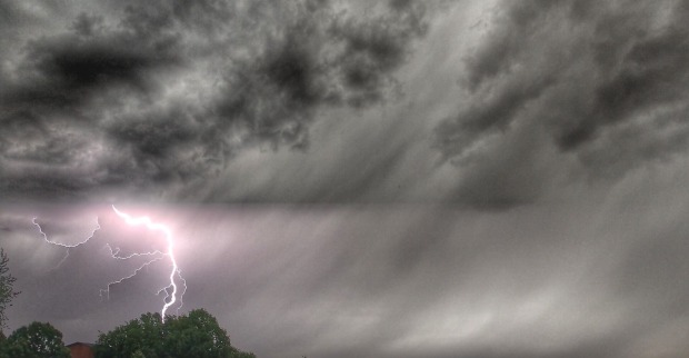 Ein heller Blitz schlägt während eines schweren Gewitters neben Bäumen ein. Der Himmel ist von dunklen, dramatischen Wolken bedeckt.