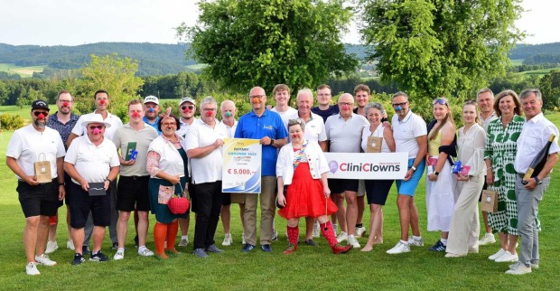 Gruppenfoto auf einem Golfplatz: Menschen mit roten Clownnasen halten einen 5.000 € Spendenscheck und ein CliniClowns-Schild.