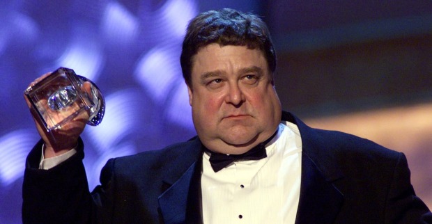John Goodman früher stark übergewichtig im Smoking, mit ernstem Blick und Pokal in der Hand.