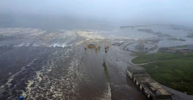 Luftaufnahme einer überfluteten Küstenstadt bei starkem Wellengang: Eine breite Tsunami-Welle überschwemmt Straßen, Häuser und Hafenanlagen unter grauem Himmel.