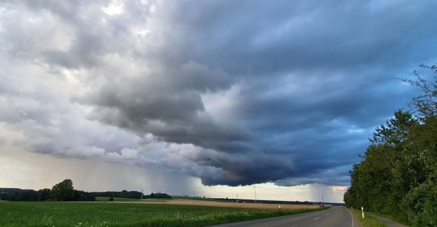 Dunkle, tief hängende Gewitterwolken über einer Landstraße und grünen Feldern, im Hintergrund ist Regen sichtbar, der aus den Wolken fällt.Bedrohliche Wolkenfront zieht über das Land – Österreich muss sich erneut auf Gewitter, Starkregen und kühle Temperaturen einstellen.