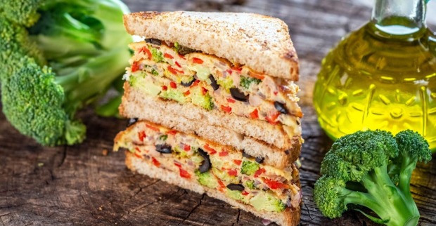 Nahaufnahme eines gestapelten Brokkoli-Sandwiches mit Paprika, Zwiebeln und Oliven, daneben frischer Brokkoli und eine Flasche Olivenöl auf Holztisch.