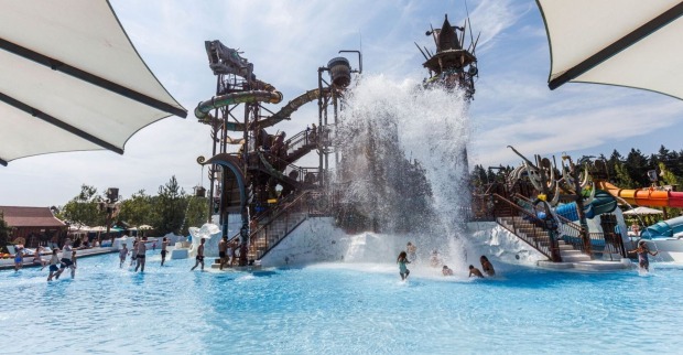 Das Bild zeigt einen großen Außenbereich eines Wasserparks mit mehreren Wasserrutschen und Spielanlagen im Hintergrund. Im Vordergrund befindet sich ein großes Becken, in dem gerade eine große Wassermenge aus einem Kipp-Eimer herabstürzt. Mehrere Kinder und Erwachsene spielen und baden im Wasser. Über dem Becken spenden weiße Sonnensegel Schatten. Im Hintergrund sind bunte Röhrenrutschen und weitere Wasserattraktionen zu sehen