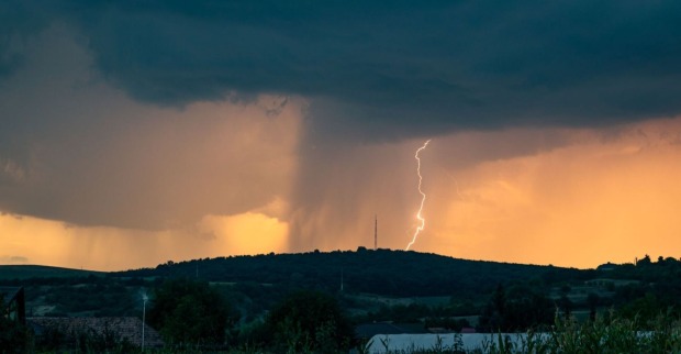 Dunkle Gewitterwolken über einer Hügellandschaft, ein Blitz schlägt neben einem Funkturm ein, im Vordergrund sind Häuser und Felder zu sehen.