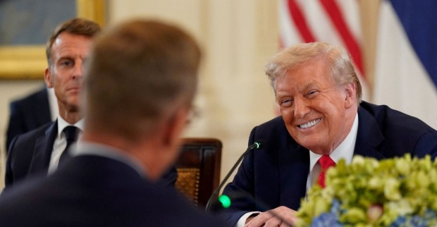 Donald Trump lächelt breit bei einem Treffen im Weißen Haus,  im Hintergrund Emmanuel Macron mit ernstem Blick.