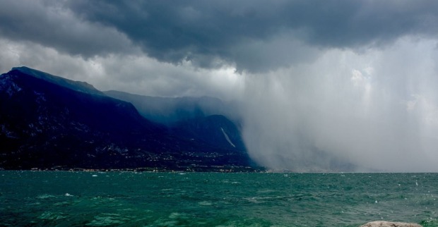 Dunkle Gewitterwolken und heftiger Starkregen über dem Gardasee. Der linke Bildteil zeigt dunkle Berge unter einer bedrohlichen Wolkendecke, rechts fällt dichter Regen über die Küste. Im Vordergrund ist aufgewühltes, türkisgrünes Wasser zu sehen.