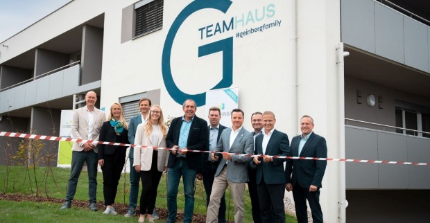 Zu sehen ist das neue Teamhaus für die Mitarbeiter des Spa Resort Geinberg. Im Vordergrund eine Gruppe von Menschen, die sich über die Eröffnung freuen. Einer der Anwesenden durschneidet ein rotes Band.