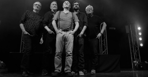 Schwarz-Weiß-Foto einer fünfköpfigen Band auf der Bühne, die nebeneinander in die Kamera blickt. Die Musiker tragen legere Kleidung und stehen vor einer Bühnenkulisse mit Scheinwerfern.