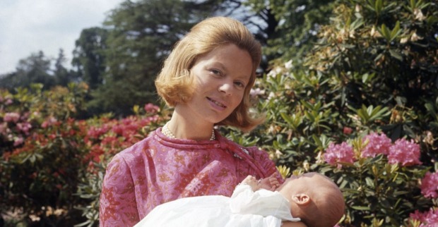Junge Frau in rosa Kleid mit Perlenkette hält ein Neugeborenes im Arm, im Hintergrund blühende Sträucher und Bäume.