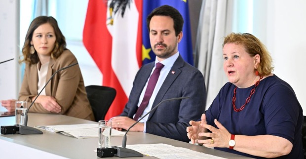 Drei Personen sitzen bei einer Pressekonferenz an einem Tisch mit Mikrofonen und Unterlagen, im Hintergrund sind österreichische und EU-Flaggen zu sehen.