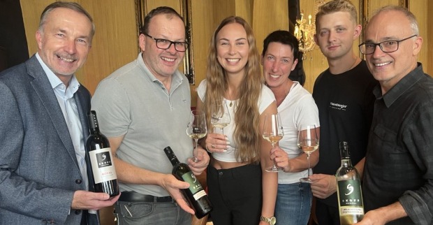 Vier Männer und zwei Männer lachen in die Kamera. In den Händen halten sie Weinflaschen beziehungsweise Weingläser, die mit Weißwein gefüllt sind.