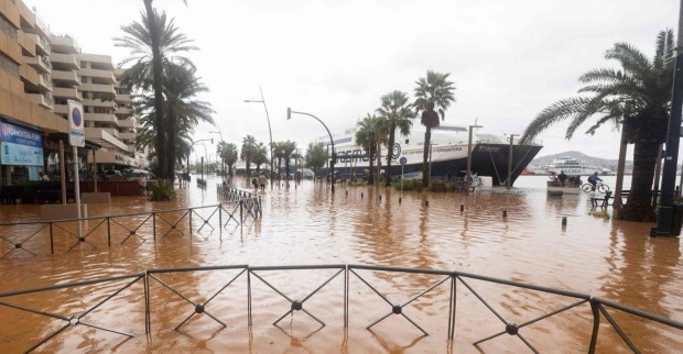 Überflutete Straßen im Hafen von Ibiza Stadt mit stehenden Wassermassen, Palmen und festliegenden Fähren im Hintergrund.