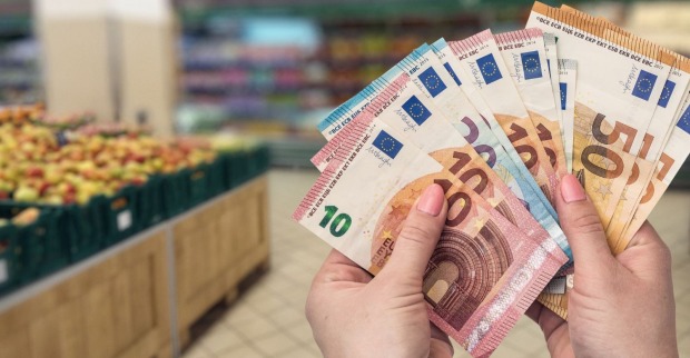 Eine Person hält mehrere Euro-Banknoten in der Hand, im Hintergrund sind Obstregale in einem Supermarkt zu sehen; Themenbild für die neuen EU-Regeln zur Behebung von Bargeld im Supermarkt.