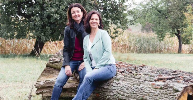 Zwei lächelnde Frauen sitzen auf einem umgestürzten Baumstamm in einer herbstlichen Wiese mit Bäumen und Maisfeld im Hintergrund.