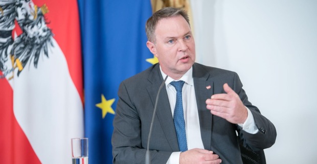 Andreas Babler in grauem Anzug gestikuliert während einer Pressekonferenz, hinter ihm sind die österreichische und die EU-Flagge zu sehen.
