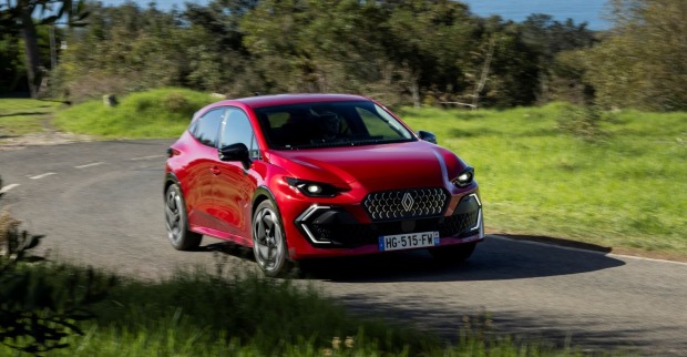 Ein roter Renault Clio fährt auf einer Landstraße. Neben der Straße ist eine grüne Wiese zu sehen, im Hintergrund ein Laubwald.