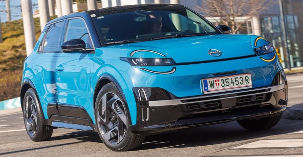 Ein hellblauer Nissan Micra glänzt in der Sonne. Das Auto parkt vor einer Glasfront, zu sehen sind auch einige Bäume.