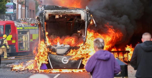 Brennender Bus bei Ausschreitungen in Belfast, Feuerwehr im Einsatz, Flammen und schwarzer Rauch auf Straße