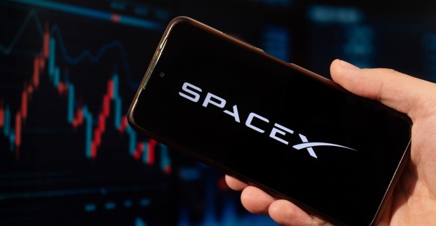 Eine männliche Hand, die ein Smartphone mit der rechten Hand hält, mit dem SpaceX Logo und einem unscharfen Börsenkonzept auf dem Hintergrund.