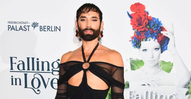 Conchita Wurst alias Tom Neuwirth bei einem Red-Carpet-Auftritt in Berlin. Die ESC-Gewinnerin trägt ein schwarzes Off-Shoulder-Outfit und posiert vor einer Fotowand.