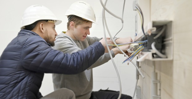 Zwei junge Personen mit weißen Schutzhelmen arbeiten gemeinsam mit Baumaterialien.