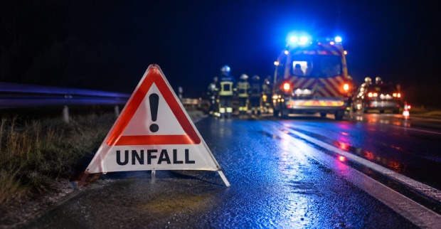 Bier-Lkw-Unfall bei Nacht mit Warndreieck und Aufschrift „Unfall“, Feuerwehrfahrzeug mit Blaulicht und Einsatzkräften auf nasser Fahrbahn.