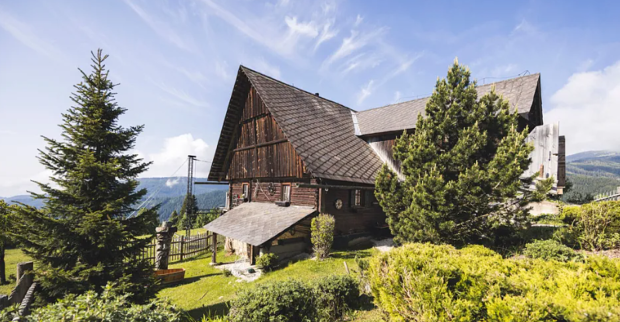 Außenansicht der Forsthaus-Rampensau-Hütte von ATV und Joyn in Kärnten, umgeben von grünen Wiesen, Tannen und einer Berglandschaft.