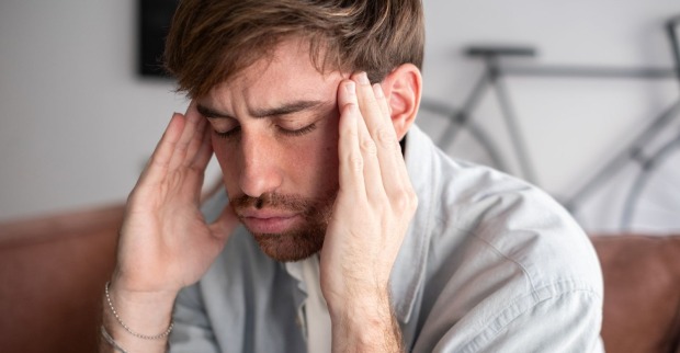 Mann sitzt mit geschlossenen Augen auf einer Couch und hält sich wegen Kopfschmerzen beide Schläfen. Kopfschmerzen können ein Symptom einer Hirnhautreizung sein.