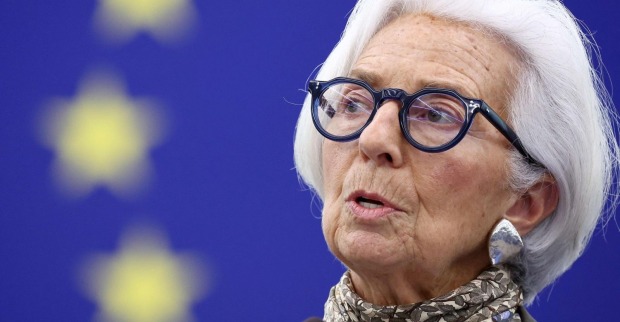 Nahaufnahme von Christine Lagarde mit weißem Haar und schwarzer Brille vor blauem Hintergrund mit unscharfen EU-Sternen.
