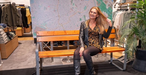 Ex-ORF-Lottofee Martina Kaiser sitzt lächelnd in einem Modegeschäft auf einer Holzbank und posiert in einem Leopardenkleid mit schwarzer Lederjacke und Stiefeln.
