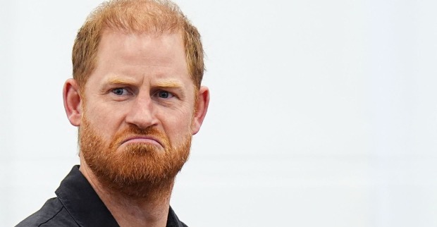 Nahaufnahme von Prinz Harry mit ernstem Gesichtsausdruck, rotem Bart und schwarzem Oberteil vor hellem Hintergrund.