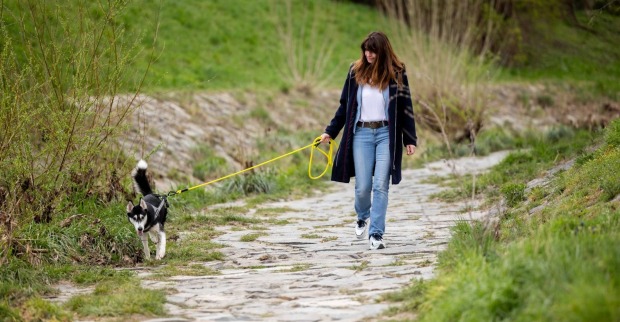 Frau in Jeans und im dunklen offenen Mantel hält einen Hund auf einem Naturweg an der Leine