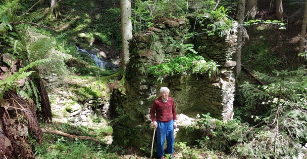 Ein alter Mann mit Gehstock steht vor der Ruine eines Partisanenunterschlupfs in einem Wald
