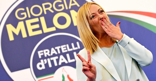 Giorgia Meloni vor einem Transparent ihrer Partei. Sie wirft eine Kusshand und zeigt das Victory-Zeichen.
