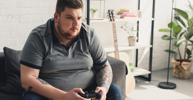 Ein übergewichtiger Mann sitzt mit dem Controller einer Spielkonsole in der Hand auf einer Couch