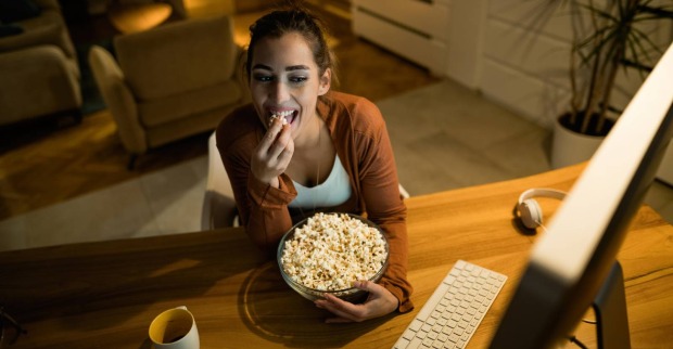 Junge Frau sieht popcorn-essend auf den Monitor ihres Computers | Credit: iStock.com/Drazen Zigic