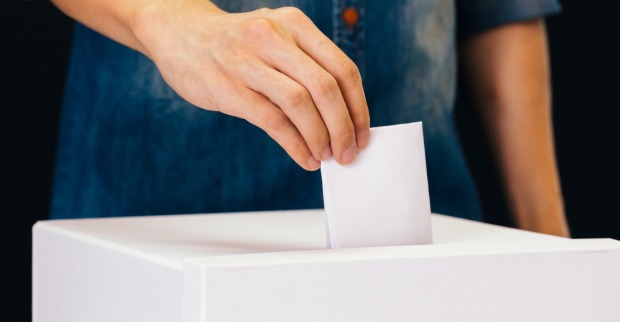 Eine Wahlurne, in die eine Person in einem Jeanshemd einen weißen Wahlzettel wirft