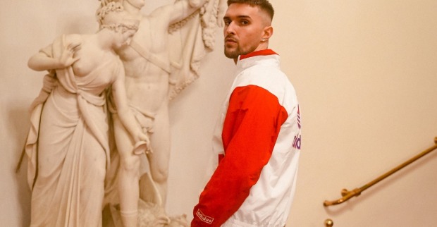Der Rapper Gavis Dean, der in eine weiße Jacke mit roten Ärmeln gekleidet, vor einer Marmorstatue steht